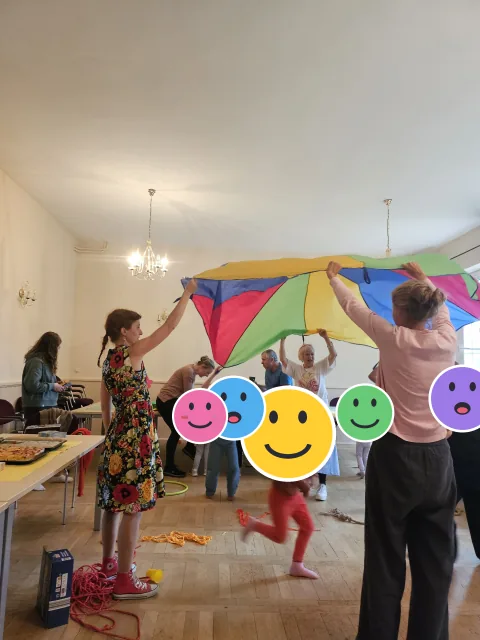 Kinder und Erwachsene spielen beim Spielplatz-Café mit einem Schwungtuch; die Kinderköpfe sind mit Smileys verdeckt